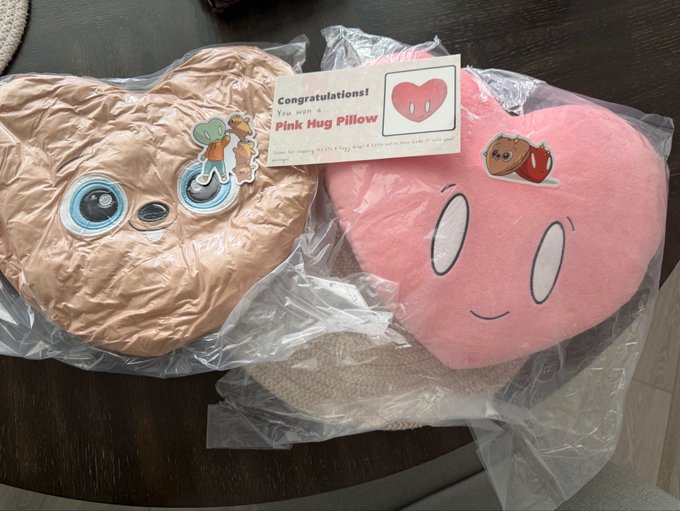 Two character-shaped pillows in plastic packaging sit on a dark surface, with a card reading 'Congratulations! You won a Pink Hug Pillow' featuring character stickers.