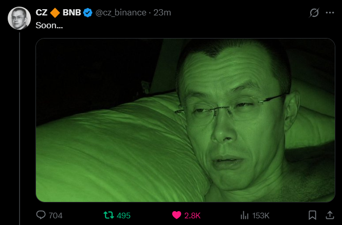 A tweet from CZ Binance (@cz_binance) posted 23 minutes ago saying 'Soon...' accompanied by a green-tinted night vision photo of a man lying in bed, with 704 replies, 495 reposts, 2.8K likes, and 153K views.