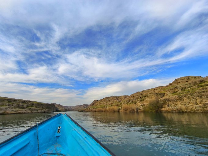A view from the bow of a bright blue boat cruising on a calm river, flanked by rocky desert hills under a vast blue sky filled with wispy clouds.