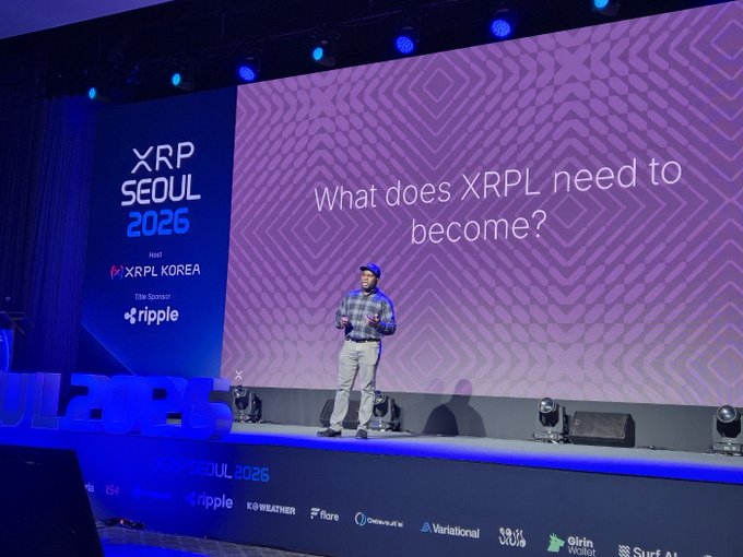 A presenter addresses attendees at the XRP Seoul 2026 conference, standing on stage beneath a large screen displaying the prompt "What does XRPL need to become?" The image captures a pivotal moment of the event, visually reinforcing the post's theme of Seoul's role in shaping the next wave of XRP Ledger adoption through developer, institutional, and user-driven infrastructure.