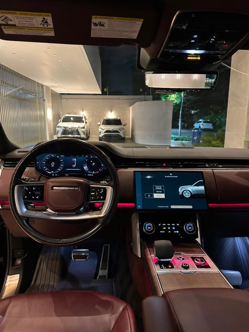 Interior view of a Range Rover, showcasing its digital dashboard, steering wheel, and central infotainment screen, with other vehicles visible in the background.