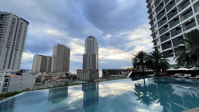 A scenic rooftop infinity pool view overlooking a tropical city skyline with high-rise buildings under a partly cloudy sky at dusk, reflecting the clouds and palm trees in the water.