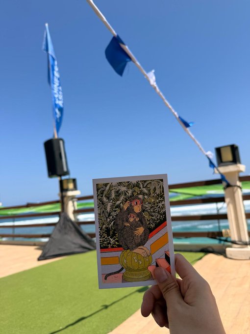 A hand holds up a colorful flyer featuring an illustrated figure wearing sunglasses and a crown, with a coin symbol, against a sunny beachside venue with a blue "Coinfest Asia" flag waving overhead—matching the caption's mention of running out of flyers at the Coinfest Asia event.