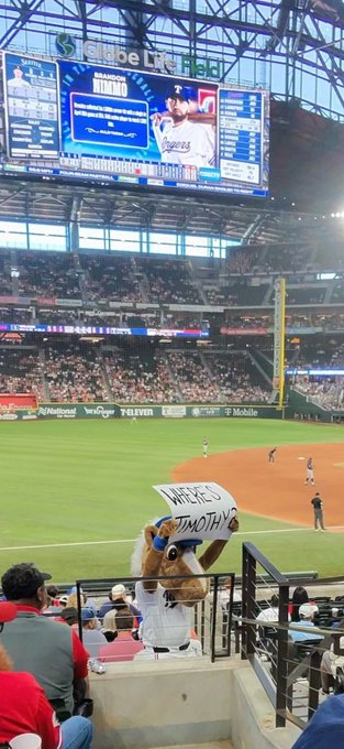 A photo taken from the stands of a baseball game at Globe Life Field, showing the Texas Rangers on the jumbotron, with a mascot in the foreground holding a sign that reads 'WIPE OUT TIMOTHY'.