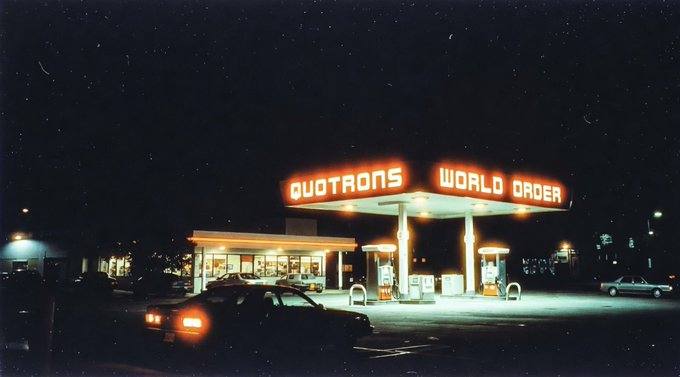 A nighttime image of a gas station with illuminated neon signs reading "QUOTRONS" and "WORLD ORDER".