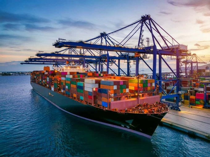 A large container ship loaded with colorful shipping containers is docked at a port with blue cranes under a sunset sky.