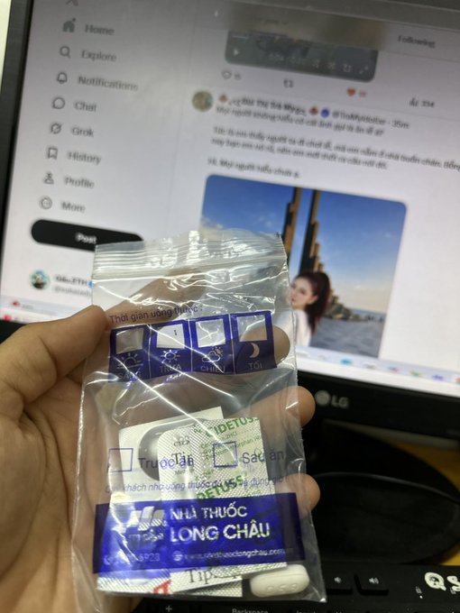 A hand holding a medication package from 'NHÀ THUỐC LONG CHÂU' (Long Chau Pharmacy) with usage instructions in Vietnamese, in front of a computer screen showing Twitter/X.