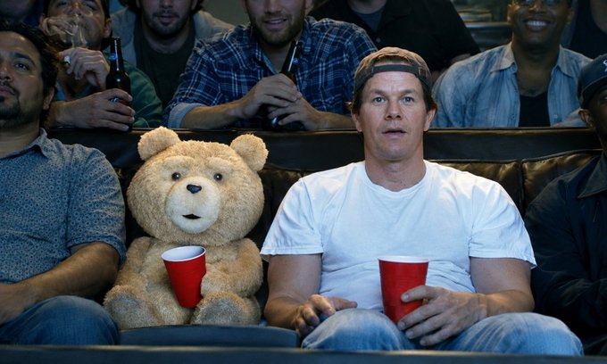 Movie still showing Mark Wahlberg sitting next to the animated character Ted the bear at a sports game, both holding red Solo cups.