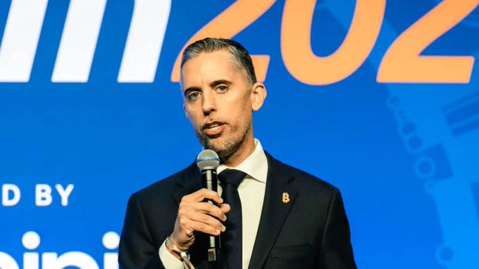 A man in a dark suit and tie speaks into a microphone at what appears to be the Bitcoin 2022 conference, with the partial text "ED BY" and "20" visible on the blue backdrop behind him. The image accompanies news of Strive CEO Matt Cole's announcement to purchase an additional 1,100 Bitcoin, growing the firm's treasury holdings beyond 24,000 BTC.