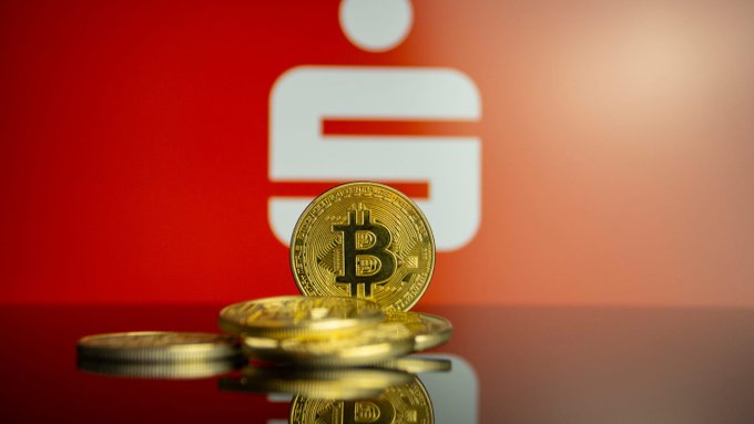 A physical Bitcoin coin stands upright on a reflective surface with blurred gold coins in front, set against a red background featuring the Sparkasse 'S' logo.