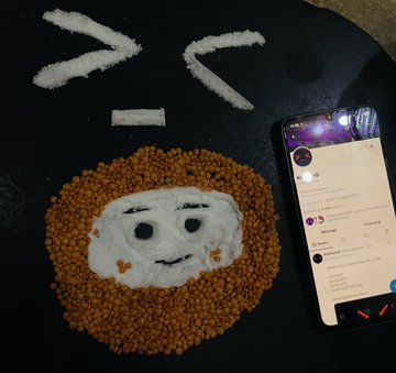 A creative arrangement of food on a black surface forms a cartoon face, with white rice for the hair and mouth and orange baked beans creating a circular border around the face, next to a smartphone displaying a social media feed.