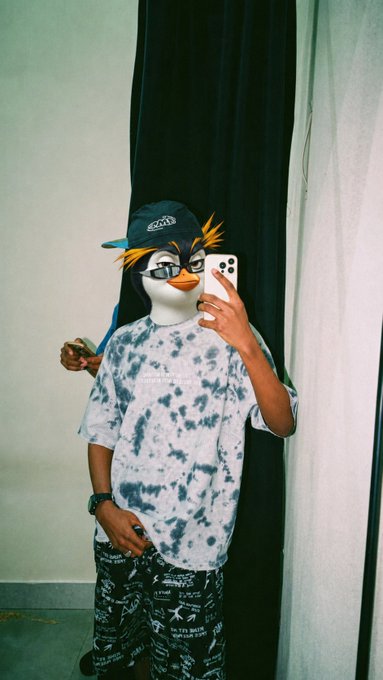 A mirror selfie of a person wearing an animated duck face filter, a black baseball cap with yellow spiky hair sticking out, and a white and grey tie-dye t-shirt with patterned shorts.
