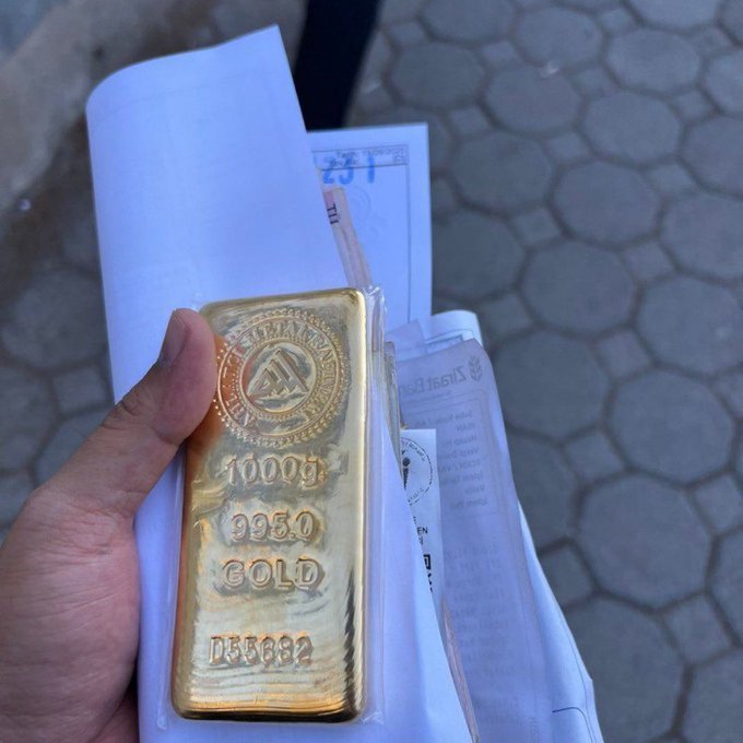 The image features a person's hand holding a large gold bar, which appears to be worth more than $1 million. The bar has various markings on its surface, indicating its value or authenticity. Next to the bar, there are several papers, one of which says "Evini satıp altın alan adamın zararı artmaya devam ediyor", which translates to "the gold is real". The presence of this information and the overall size of the gold bar convey its value or importance.