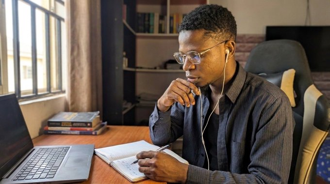 A young man wearing glasses and earphones sits at a desk by a window, thoughtfully looking at a laptop screen with a notebook and pen in hand.