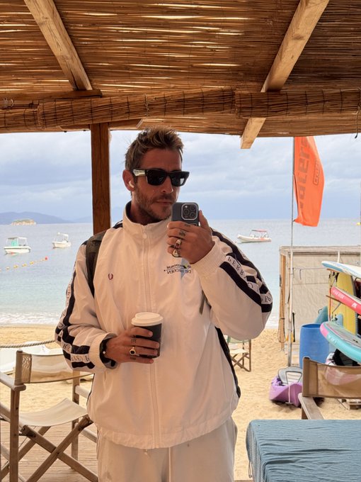 A man wearing sunglasses and a white tracksuit takes a mirror selfie on a beach while holding a coffee cup, with small boats and an orange flag in the background.