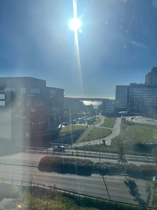 A view from a window on a sunny day showing a building, a curved road with greenery, and a body of water in the distance.