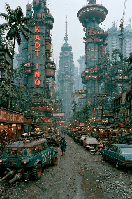 A bustling cyberpunk cityscape featuring massive industrial towers with glowing signage reading "KADTINO," crowded streets lined with weathered vehicles, and lush vegetation overtaking the decaying urban environment.