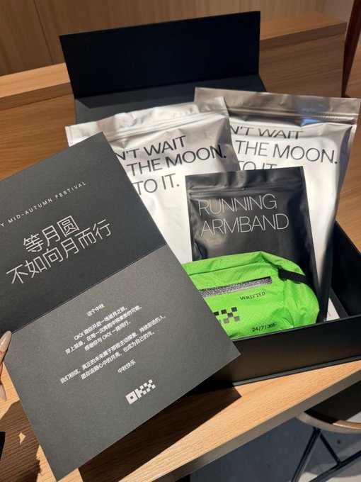 An unboxed OKX promotional gift set for a Mid-Autumn Festival, containing a greeting card with Chinese text reading '等月圆 不如向月而行', along with silver 'DON'T WAIT FOR THE MOON. GO TO IT.' packages and a green OKX running armband.
