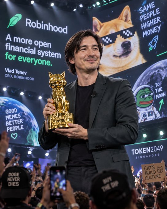 A man holding a golden Dogecoin trophy stands before a crowd, with a Robinhood backdrop displaying 'Vlad Tenev CEO, ROBINHOOD' and slogans like 'SAME PEOPLE HIGHER MONEY' and 'GOOD MEMES BETTER PEOPLE' at the TOKEN2049 Singapore event.