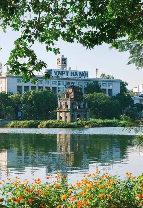 A scenic view of Hoan Kiem Lake in Hanoi, Vietnam, featuring the iconic Turtle Tower on a small island, the VNPT Hà Nội building in the background, and orange marigold flowers framing the foreground.