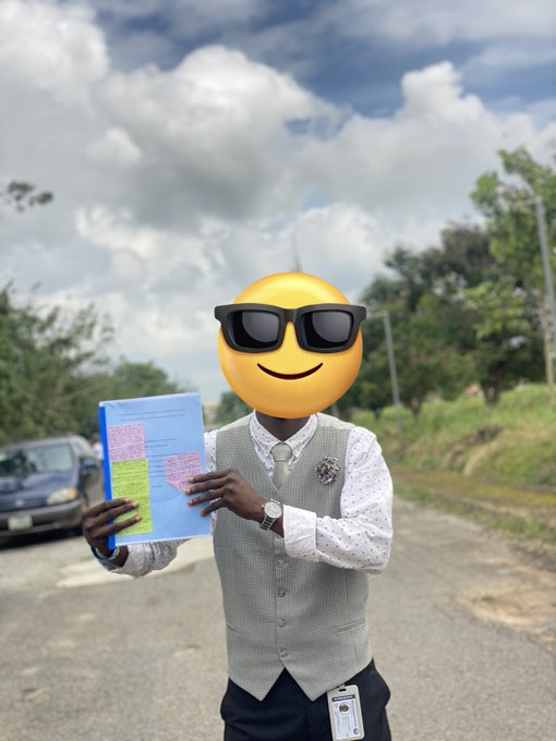 A man wearing a grey vest and white patterned shirt holding colorful papers outdoors, with a large sunglasses emoji covering his face and an ID badge clipped to his vest.