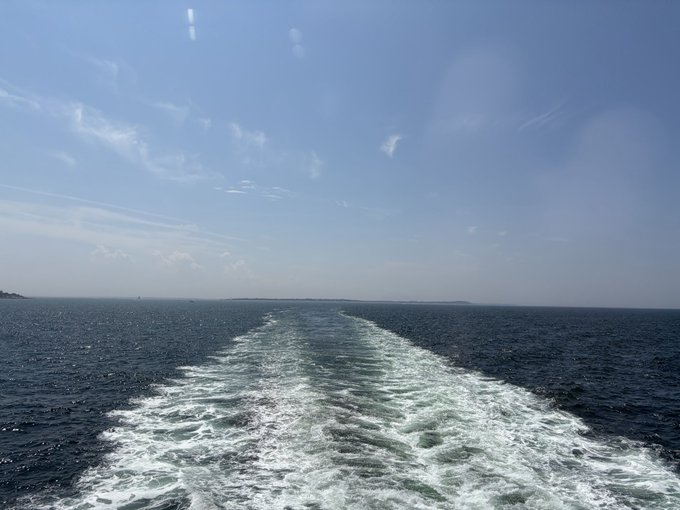 The image shows the foamy white wake of a ferry stretching across deep blue ocean waters under a bright sky, with a faint glimpse of land (Long Island) receding in the distance — perfectly capturing the "bye bye" farewell moment as the boat departs. No text, numbers, or tickers are visible in the image.