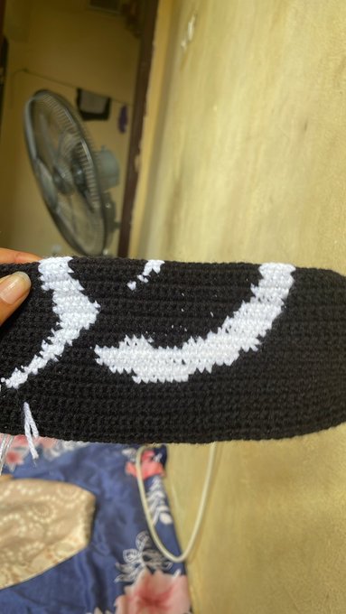 A close-up of a hand holding a black and white crocheted fabric piece featuring a crescent moon design, with a fan visible in the background.