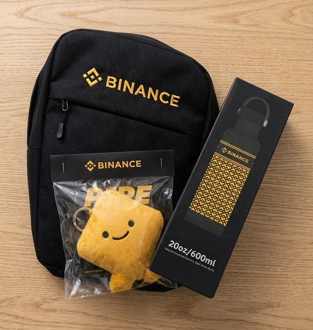 Binance promotional merchandise including a black backpack, a plush yellow mascot toy in branded packaging, and a 20oz/600ml vacuum insulated stainless steel water bottle, all displayed on a wooden surface.