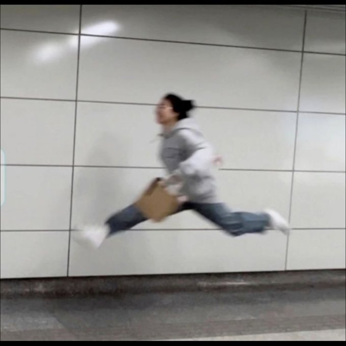 A blurry action shot of a person in a gray hoodie and jeans leaping mid-air while holding a brown object, moving past a white tiled wall.