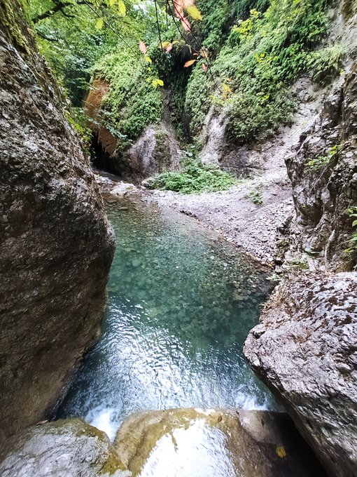 A photograph of a natural pool of clear turquoise water surrounded by moss-covered rock walls and lush greenery, with a small waterfall cascading in.