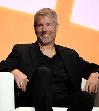 A smiling man with a gray beard wearing a black shirt and jacket, seated in a white chair.