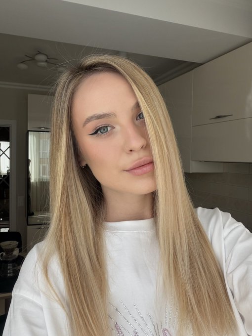 A close-up selfie of a young woman with long, straight blonde hair, wearing a white sweatshirt and blue eyeliner, standing in a kitchen with white cabinets.