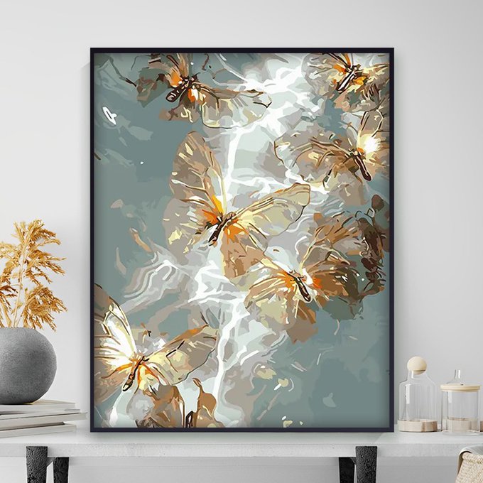 A framed painting featuring abstract golden butterflies and dragonfly-like insects against a muted teal and grey background is displayed on a white shelf alongside decorative dried plants and glass jars.