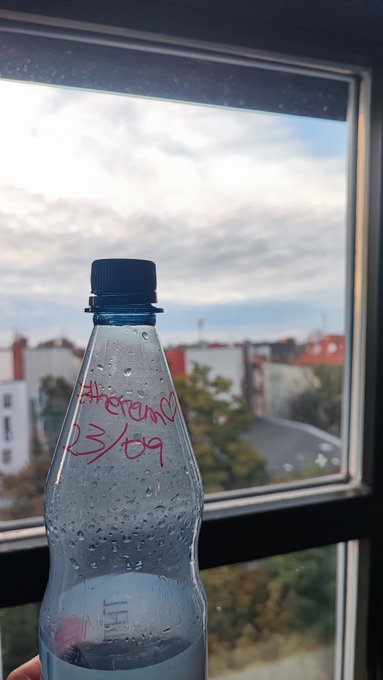 An empty clear plastic water bottle held up against a window with a city view, labeled in pink marker with "therand 23/09".