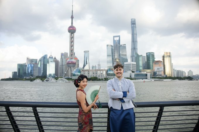 A couple posing in front of the Shanghai skyline, with the woman in a red cheongsam holding a green fan and the man in a blue and white outfit.