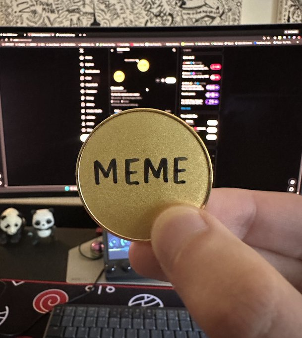 A hand holds up a gold-painted coin with "MEME" written in black Sharpie in front of a computer monitor displaying cryptocurrency charts, illustrating the creator's DIY attempt at making the first IRL memecoin ($MEME).