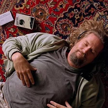 A scene of a bearded man in a gray t-shirt and green hoodie lying asleep on an ornate red Persian rug next to a smartphone and a remote control, recognizable as the character 'The Dude' from The Big Lebowski.