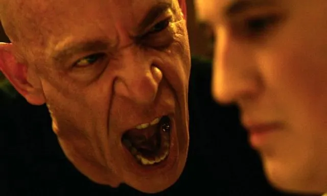 A close-up of a man with a shaved head and pointed ears yelling aggressively, while a younger person stands calmly in the background.
