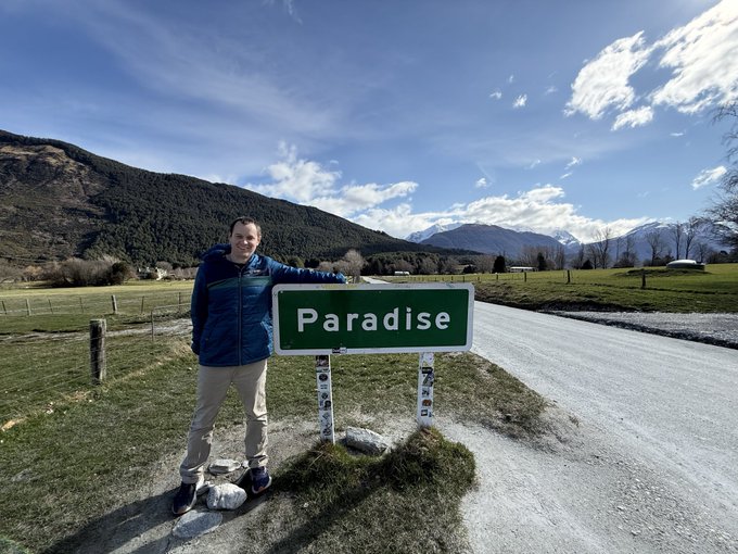 A man stands next to a green road sign reading "Paradise" in a scenic mountainous landscape, posing for a photo in what appears to be the New Zealand countryside, fitting the caption's Middle Earth reference. The Bitcoin dominance comment is humorously tied to the "Paradise" sign, suggesting a whimsical escape from the crypto markets.
