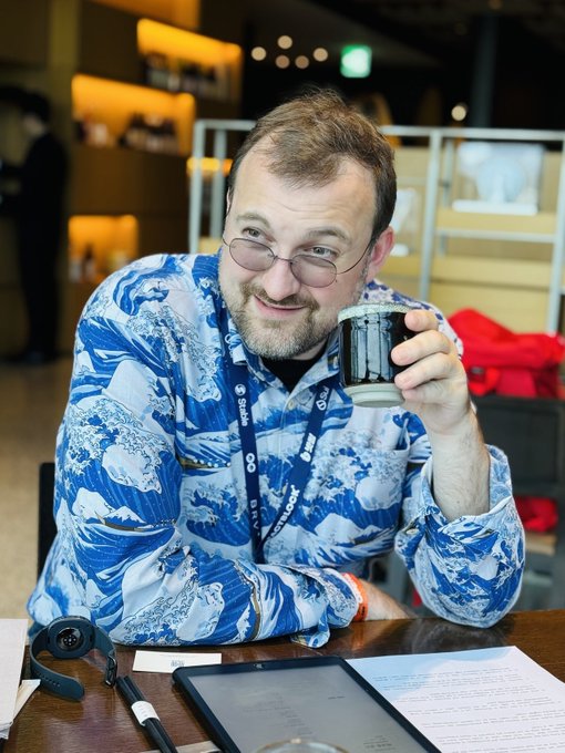 Cardano founder Charles Hoskinson, wearing a "SUNDE" lanyard and a blue wave-patterned shirt, smiles while holding a dark mug at a conference table with a tablet and headphones, illustrating the tech event context of his prediction that crypto will "eat" AI over the next 5-10 years.