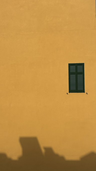 A minimalist photograph shows a green shuttered window against a textured yellow wall, with the shadow of rooftops cast along the bottom edge.