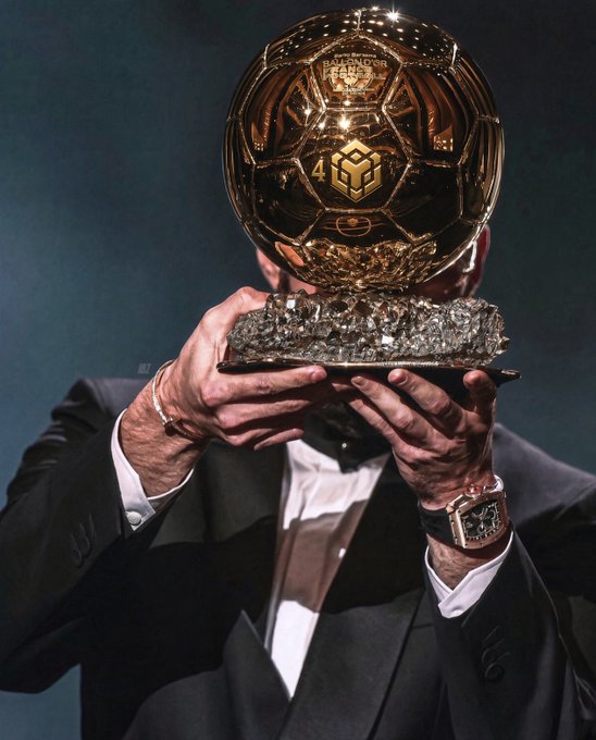 A person in a black suit holds up a golden Ballon d'Or trophy mounted on a crystal base, with a luxury watch visible on their wrist.