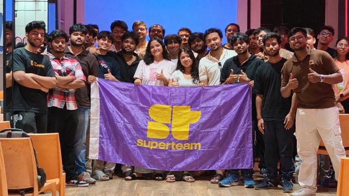 A group photo of the "superteam" community holding a purple banner with the yellow Superteam logo, taken indoors.
