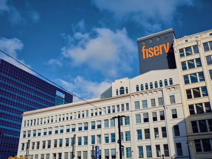 An exterior view of urban architecture under a blue sky, featuring a historic building in the foreground and a modern skyscraper displaying the orange 'fiserv' logo.