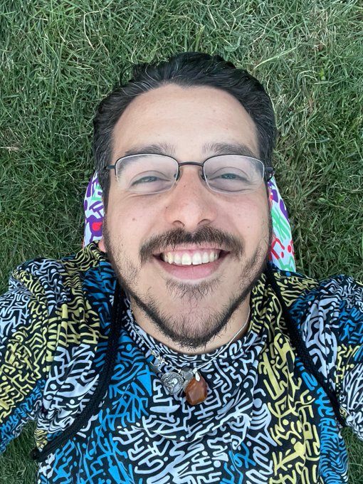A selfie of the same man from the previous photo, now lying on green grass wearing glasses and the same colorful patterned shirt, smiling at the camera.