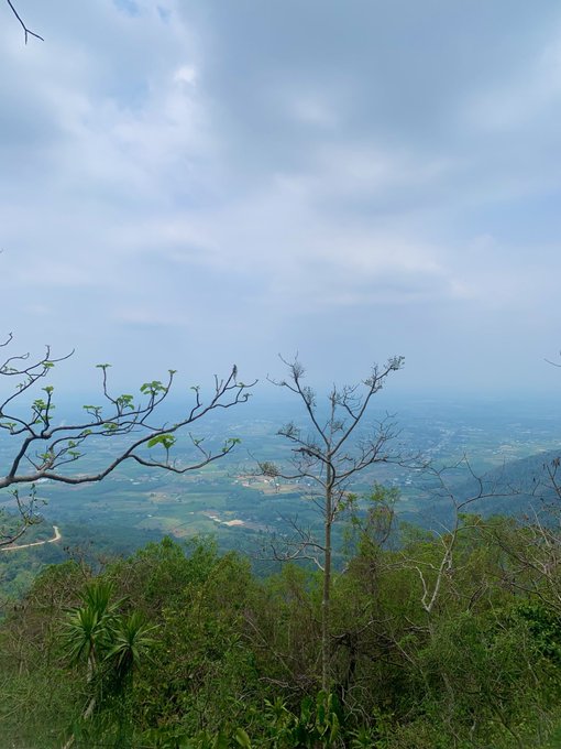 A scenic view from a mountain overlook shows a vast landscape with green hills, distant villages, and a partly cloudy blue sky, framed by sparse tree branches in the foreground.