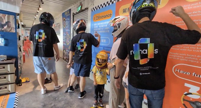 A group of people wearing black 'run as' branded t-shirts and helmets are gathered indoors, with a child in a yellow helmet being prepared, possibly at an indoor karting venue with 'Entrance' signage visible.