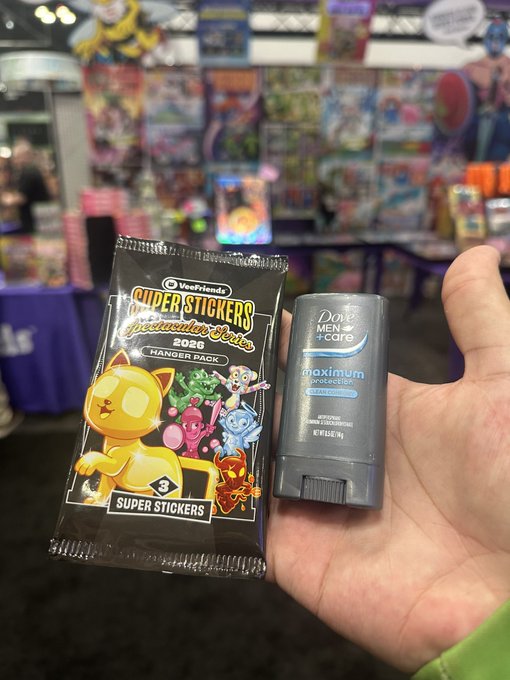 A hand holding two items at what appears to be a convention or store: a 'Super Stickers Spectacular Series 2026 Hanger Pack' by Upper Deck featuring cat-like characters, and a Dove Men+Care Maximum Protection deodorant stick.