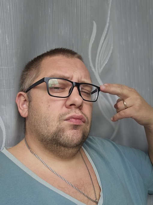 A close-up of a man with a short buzz cut and beard, wearing black-rimmed glasses and a teal v-neck shirt, touching the frame of his glasses with his finger.
