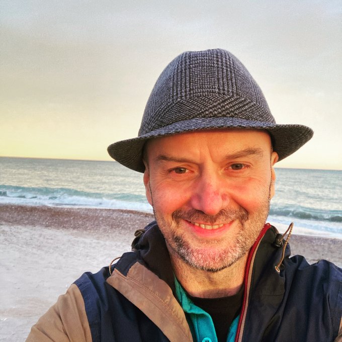 A selfie of a smiling middle-aged man with a beard wearing a gray fedora and a tan/blue jacket, standing on a beach with the ocean and a sunset sky in the background.
